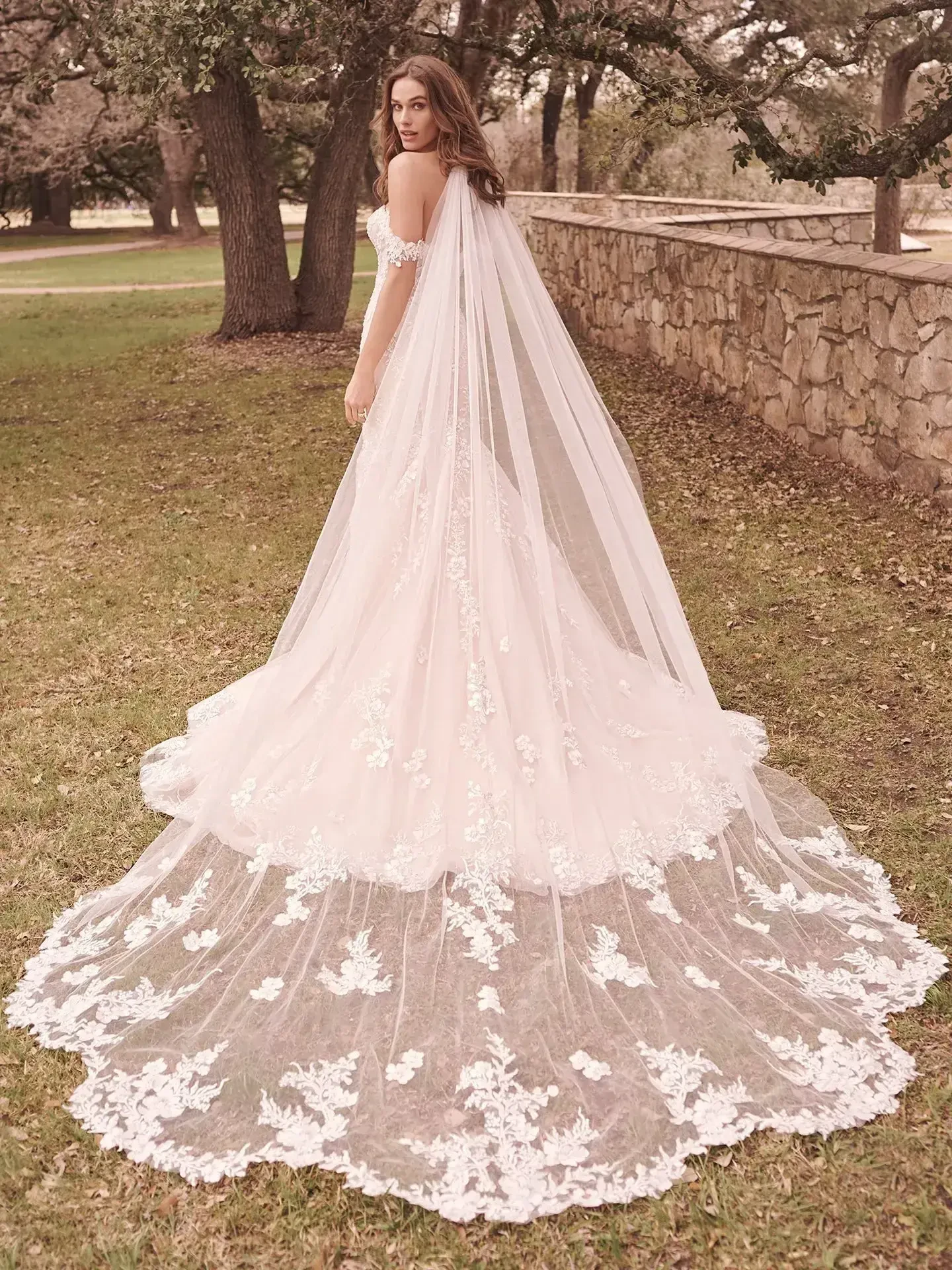 A woman in a flowing wedding dress with intricate lace details and a long train, standing outdoors in a grassy area with trees in the background.
