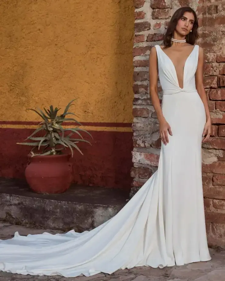 Model is wearing a sleek, fitted sleeveless wedding dress with a plunging neckline and open back.