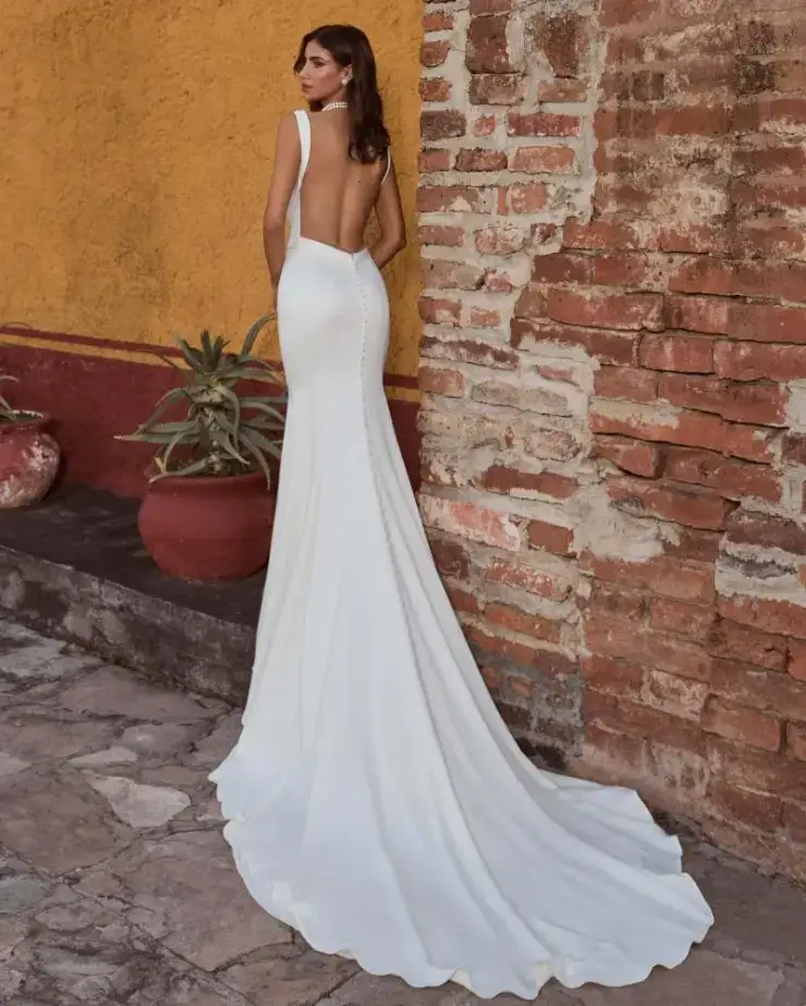 Model is wearing a sleek, fitted sleeveless wedding dress with a plunging neckline and open back.