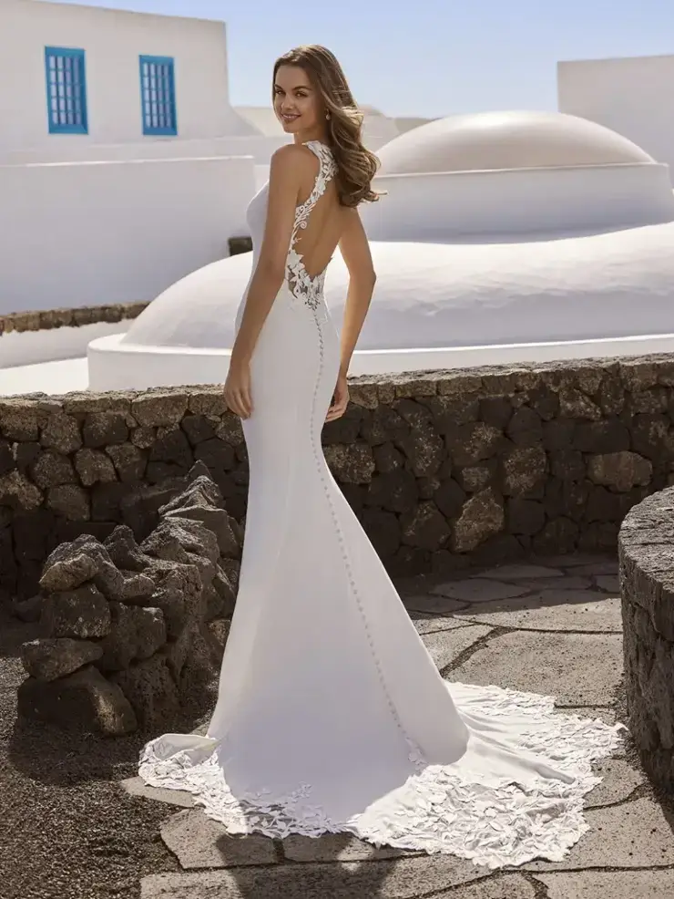 A woman in a sleek white wedding dress with lace detailing stands smiling against a backdrop of stone walls and whitewashed buildings under a clear blue sky.