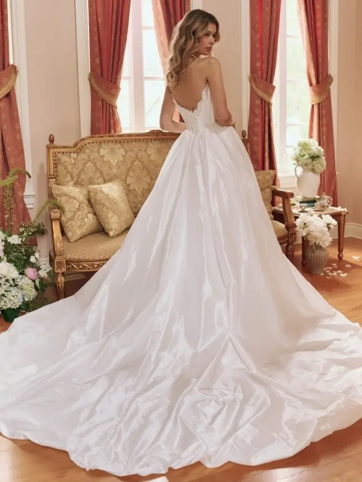 A woman in an elegant strapless wedding gown with a flowing train stands in a vintage-style room. The atmosphere is romantic and refined.