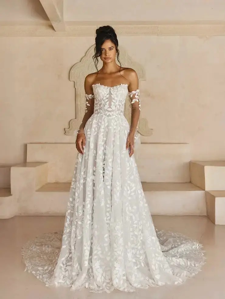 Model is wearing a strapless a-line wedding gown with detachable off the shoulder sleeves and a leaf lace pattern 