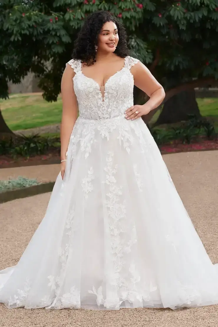 A woman wearing a beautiful white wedding gown with intricate lace details, standing outdoors in a serene setting.