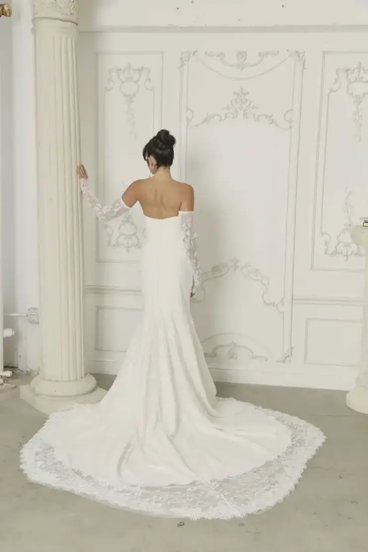 Model is wearing a strapless minimalist fit-and-flare wedding dress with detachable off-the-shoulder long lace sleeves.