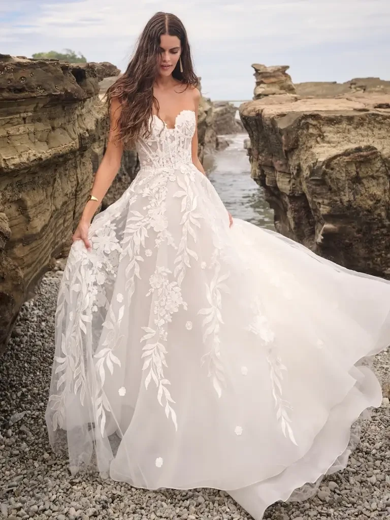 A woman in a flowing white wedding dress with floral embellishments stands on rocky terrain near the water.