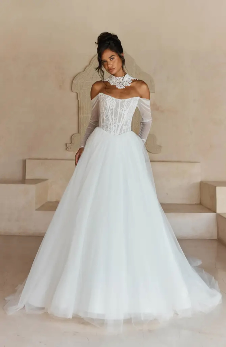 Bride in an elegant off-the-shoulder wedding gown with a full tulle skirt and lace detailing, standing against a neutral backdrop, exuding grace.