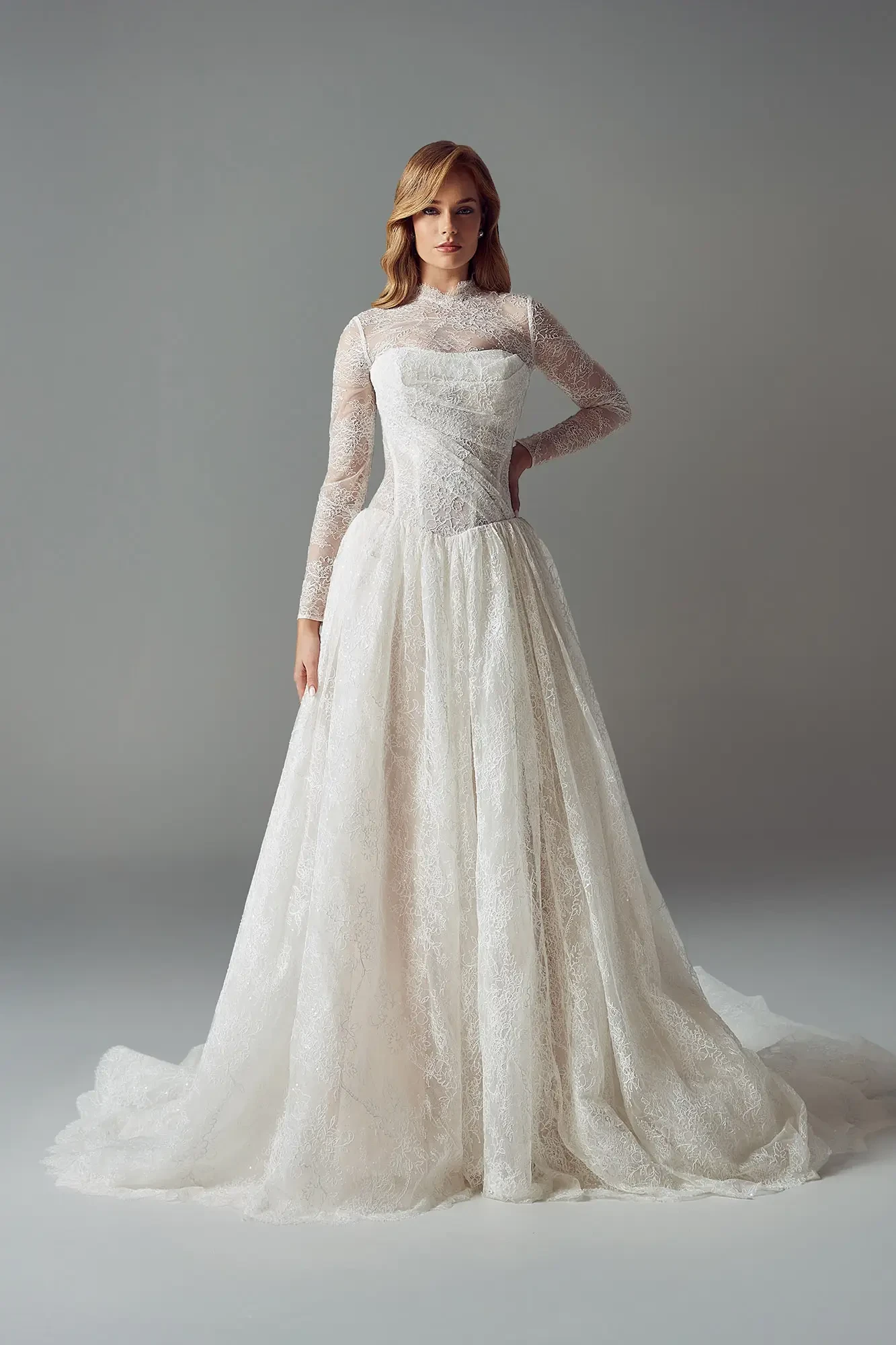 A woman in a lace wedding gown with long sleeves and a full skirt, standing confidently against a gray background, creating an elegant and serene tone.