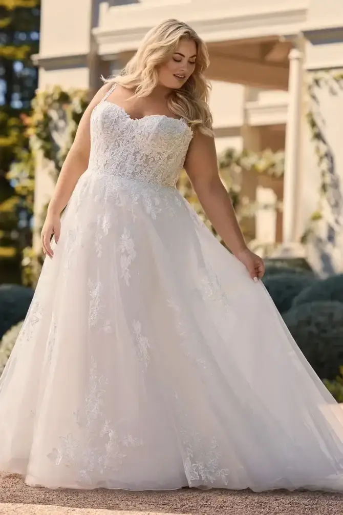 A woman in a wedding dress with lace details, standing outdoors and looking down at the fabric of her dress.