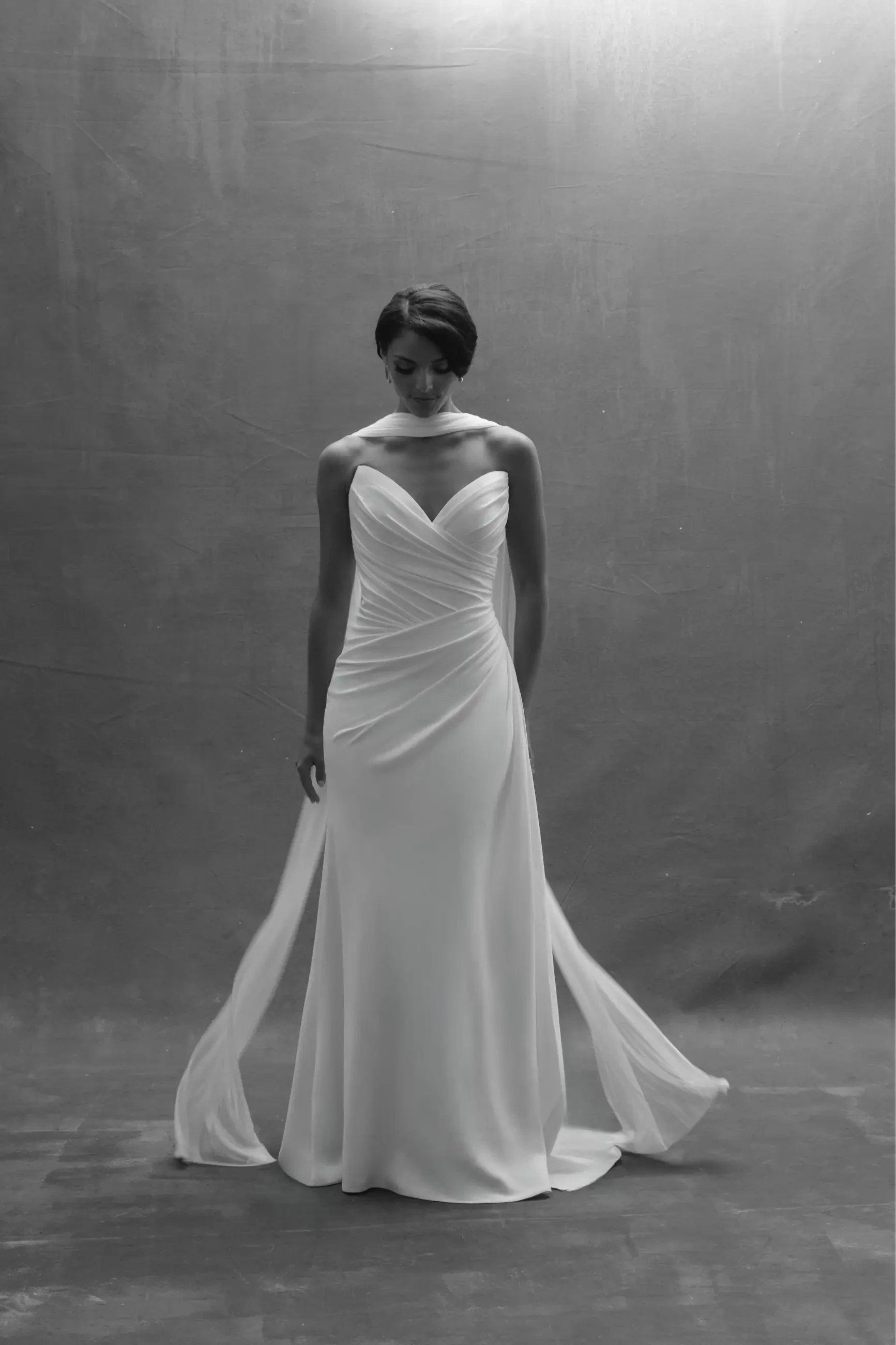 Elegant black-and-white photo of a woman in a strapless, flowing white gown. She stands gracefully on a textured background, conveying a serene, contemplative mood.