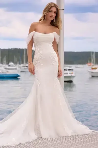 A woman in an elegant off-the-shoulder wedding dress with floral embroidery, standing on a dock with boats in the background.
