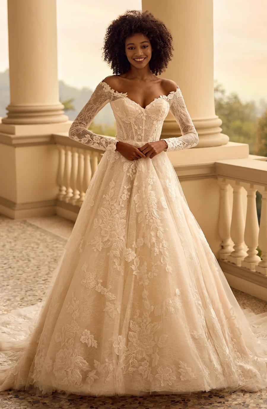 Bride in an elegant, off-shoulder lace wedding gown with long sleeves, standing on a stone balcony. The setting is serene with soft lighting.