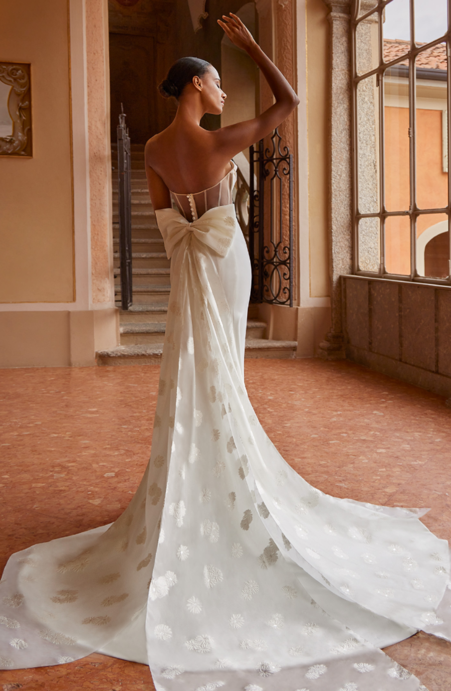 Elegant woman in a flowing white gown with a large bow, standing gracefully indoors. Sunlight streams through a large window, creating a serene ambiance.