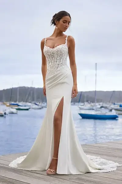 A woman in a strapless white wedding dress stands outdoors, with a textured wall and a blue window in the background.
